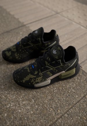 Czarne i oliwkowozielone sneakersy Adidas w kamuflażowy wzór z grubymi podeszwami, spoczywające na teksturowanych kostkach brukowych.