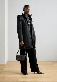 Black puffer coat with a zip front and hood, textured black handbag, wide-leg black trousers, and pointed black heels on a wooden floor.