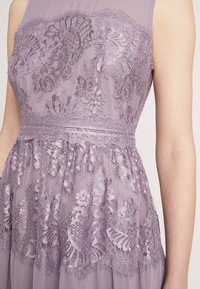 Lila spetsklänning med blommönster, genomskinlig överläggning och en strukturerad midja. Har en figursydd överdel och en svävande kjol.