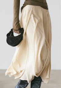 Beige vloeiende rok van lichtgewicht stof, met plooien en een brede vorm. In één hand wordt een zwarte suède handtas gedragen.