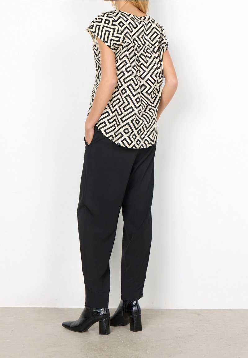 Femme portant un haut à manches courtes à motif géométrique noir et blanc, un pantalon noir et des bottines à talons noires, debout contre un mur blanc.