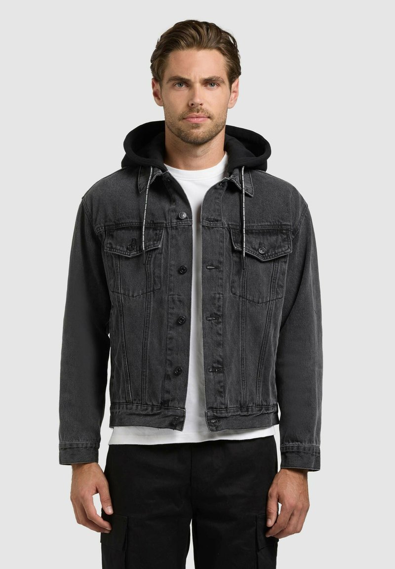 Homme portant une veste en denim noire avec capuche noire sur une chemise blanche, debout devant un fond gris clair uni.