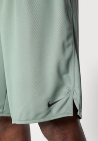 Grüne Sportshorts aus atmungsaktivem Material mit strukturiertem Muster, ausgestattet mit einem schwarzen Nike-Logo am linken Saum und seitlichen Schlitzen.