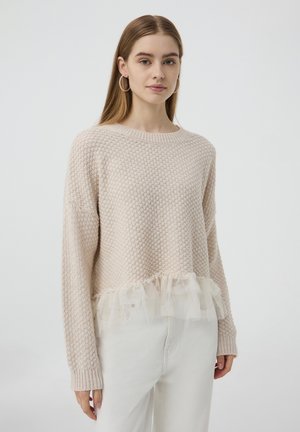 Maglione beige chiaro con un design a maglia testurizzata, caratterizzato da un orlo in tulle arricciato. Maniche lunghe e scollatura rotonda. Indossato sopra jeans bianchi.