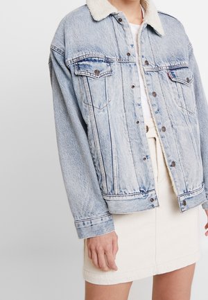 Veste en denim bleu clair avec col en polaire blanche portée sur une jupe et un top blancs sur une personne devant un fond uni.