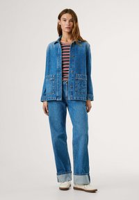 Denimjas met twee voorzakken, blauw van kleur, gedragen over een gestreept rood-wit shirt. Gecombineerd met wijdlijnige blauwe spijkerbroeken en witte schoenen.