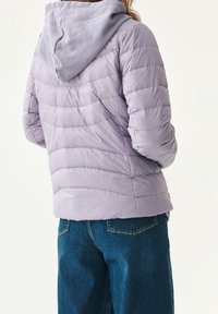 Lilla polstret jakke med hætte, prydet med et quiltet design og elastiske manchetter. Bæres over blå jeans, som fremhæver et afslappet, lagdelt look.