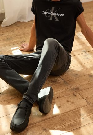 Osoba siedząca na drewnianej podłodze, ubrana w czarną koszulkę Calvin Klein Jeans, ciemnoszare dżinsy i czarne buty, z jedną wyprostowaną nogą.