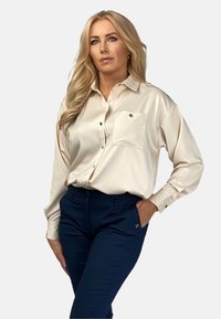 Kremowa satynowa koszula z guzikami, z jedną kieszenią na piersi, w oversizowym kroju i złotymi akcentami, zestawiona z granatowymi spodniami. Miękka tekstura.