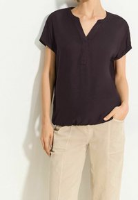 Blouse courte à manches courtes couleur violet foncé avec un décolleté en V et une coupe décontractée, associée à un pantalon beige clair avec poches. Texture de tissu lisse.