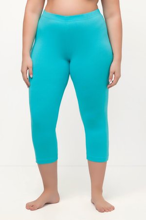 Frau trägt enganliegende türkisfarbene kurze Leggings, steht barfuß vor einem schlichten weißen Hintergrund.