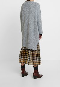 Long gray knitted cardigan with a textured pattern, side slits, paired with a plaid tiered skirt in yellow and black, and brown heeled boots.