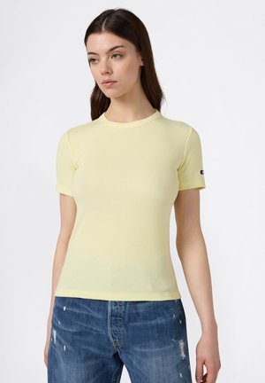 Jeune femme aux longs cheveux bruns portant un top côtelé à manches courtes jaune clair et un jean bleu déchiré, debout devant un fond blanc.