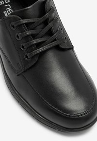 Chaussure en cuir noir avec une finition lisse. Présente un bout arrondi, six œillets et des lacets noirs plats. Accents cousus le long des côtés.