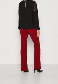 Blouse noire avec des manches en dentelle, détail en trou de serrure au dos, associée à un pantalon rouge ajusté et des chaussures à talons noirs. Texture lisse, design élégant.