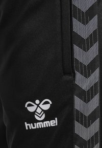 Svarta träningsoveraller med en vit Hummel-logotyp. Sidostrips har ett chevronmönster i vitt mot en texturerad bakgrund.