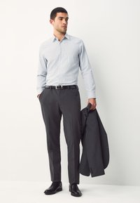 Világoskék mintás ing, szürke elegáns nadrág, fekete öv és fényes cipők. Egy sötétszürke blézert tart, sima anyagból.