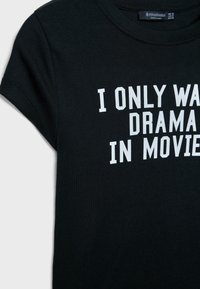 Musta puuvillapaita, jossa on valkoista tekstiä, joka lukee "HALUAN VAIN DRAMAA ELokuvissa." Taitettu kaula-aukko, lyhyet hihat ja rento istuvuus.