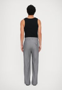 Man gezien van achteren, gekleed in een zwarte mouwloze top en grijze wijde broek, tegen een effen lichte achtergrond.