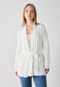 White ribbed cardigan with a tie belt, featuring two front pockets and long sleeves. Worn over a light top and blue denim.