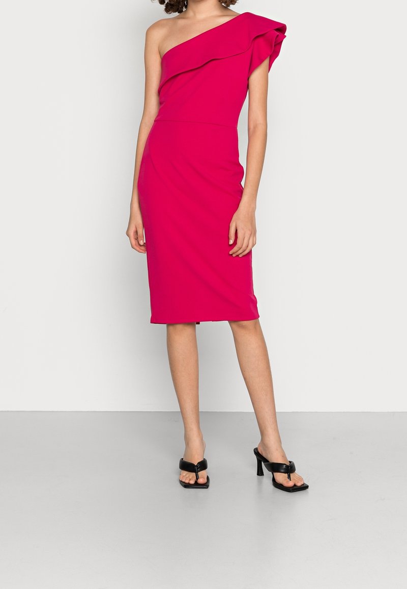 Femme portant une robe rose vif une épaule longueur genou et des sandales noires à talons ouverts, debout devant un fond uni.