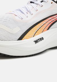 Zapatilla deportiva blanca con parte superior de malla transpirable, con franjas de acento en naranja y negro, una entresuela blanca acolchada y suela de tracción negra.