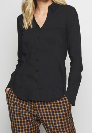 Mujer con una camisa negra ajustada de botones, con cuello y mangas largas, combinada con pantalones marrones y negros de cuadros, manos en los bolsillos.