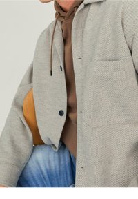 Szara marynarka w kratę pepitkę, wykonana z teksturowanej tkaniny, z kieszenią na piersi i czarnymi guzikami, noszona na brązowej bluzie z kapturem i niebieskich dżinsach.