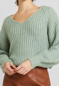 Blekgrön stickad tröja med V-ringning och ribbade ärmar, med ett strukturerat mönster. Modellen bär en brun läderkjol.