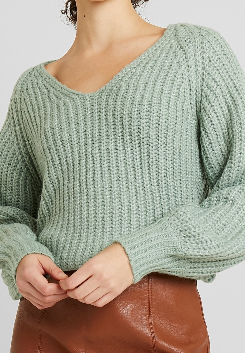 Blekgrön stickad tröja med V-ringning och ribbade ärmar, med ett strukturerat mönster. Modellen bär en brun läderkjol.