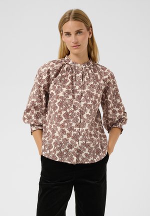 Femme blonde les mains dans les poches, portant un chemisier à manches longues à motif floral avec un col froncé et un pantalon noir.