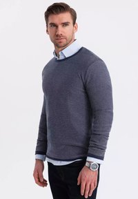 Marinblå stickad tröja med ett strukturerat mönster, rundad halsringning och kontrasterande ribbade ärmslut. Bärs över en ljusblå skjorta med knappar.