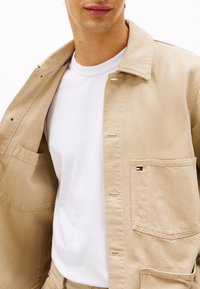 Beige Baumwolljacke mit Kragen, Vordertasche und Knopfverschluss, getragen über einem weißen T-Shirt, mit kleinem Markenlogo auf der Tasche.