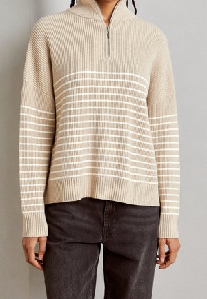 Beiger, gerippter Strickpullover mit weißen horizontalen Streifen, hoher Kragen mit Reißverschluss, getragen mit schwarzen Hosen.