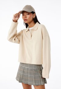 Beige langærmet polo med tre knapper, parret med en plisseret mini-skirt i grå og brun tern, samt en matchende kasket.