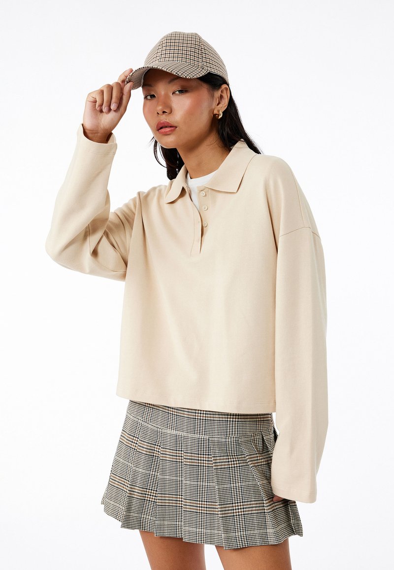 Beige langærmet polo med tre knapper, parret med en plisseret mini-skirt i grå og brun tern, samt en matchende kasket.