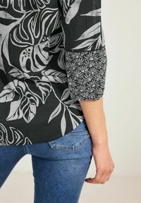 Maglietta nera con stampe botaniche bianche, dotata di un polsino a contrasto. Indossata con jeans blu, che mostrano un orlo inferiore arrotondato.