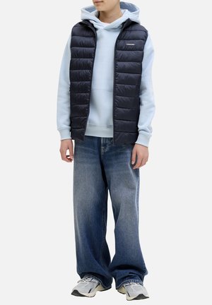 Personne portant un sweat à capuche bleu clair, un gilet matelassé noir, un jean bleu ample et des baskets blanches, debout devant un fond uni.