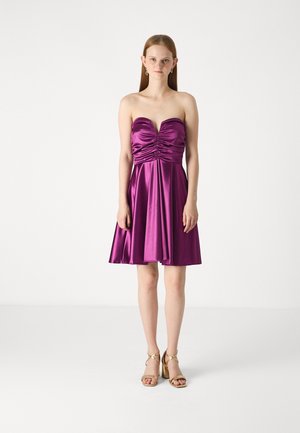 Robe en satin violet sans bretelles avec un corp en drapé, une jupe évasée et un ourlet au genou. Le modèle est associé à des sandales dorées à talons hauts.