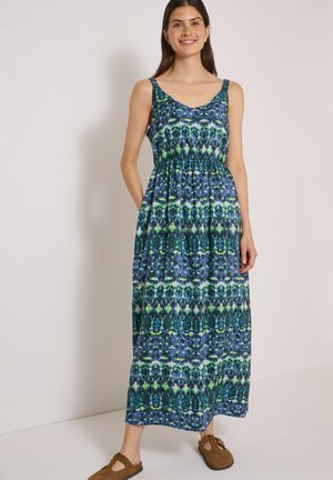 Femme souriante portant une robe maxi sans manches à motif bleu et vert avec des poches et des chaussures marron à enfiler, debout contre un mur uni.