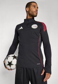 Black long-sleeve soccer training top with red accents, zip collar, and logo. Model holding a soccer ball, wearing black shorts.