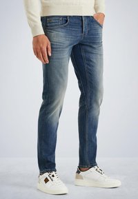 Jeans azul de corte Slim fit con un ligero desteñido, diseño de cinco bolsillos y costuras sutiles. Llevados con zapatillas blancas que presentan detalles en negro y beige.