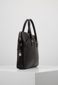Sort læder handbag med dobbelte hændler, guld lynlås og lille rund guld charm, udstillet på en hvid pedestals mod en grå baggrund.