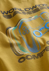 Geel katoenen t-shirt met een grafiek van een gamecontroller in blauwe en teal accenten, met daarboven de tekst "WORLDWIDE GAMER".