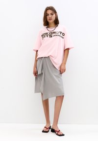 Lys pink oversized t-shirt med grafisk tekst, grå asymmetrisk indpakningsskørt og sorte kvadratiske hælsandaler med røde remme.