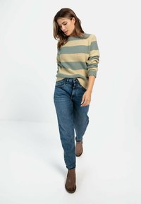 Geribbelde trui in lichtgroene en beige strepen, gecombineerd met losse blauwe jeans en bruine enkellaarsjes. Model loopt met het hoofd iets naar beneden.