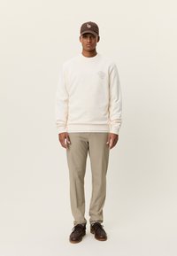 Μπλούζα φούτερ σε εκρού χρώμα με υφή ύφανσης, με τυπωμένο σχέδιο στο στήθος. Συνδυάζεται με ανοιχτό μπεζ παντελόνια και σκουρό καφέ παπούτσια.