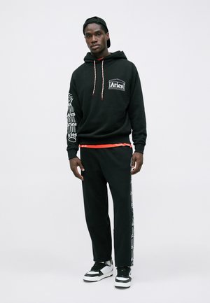 Homem a usar um moletom preto da Aries e calças combinando com gráficos em texto branco, boné preto virado para trás e sapatilhas em preto e branco, de pé.