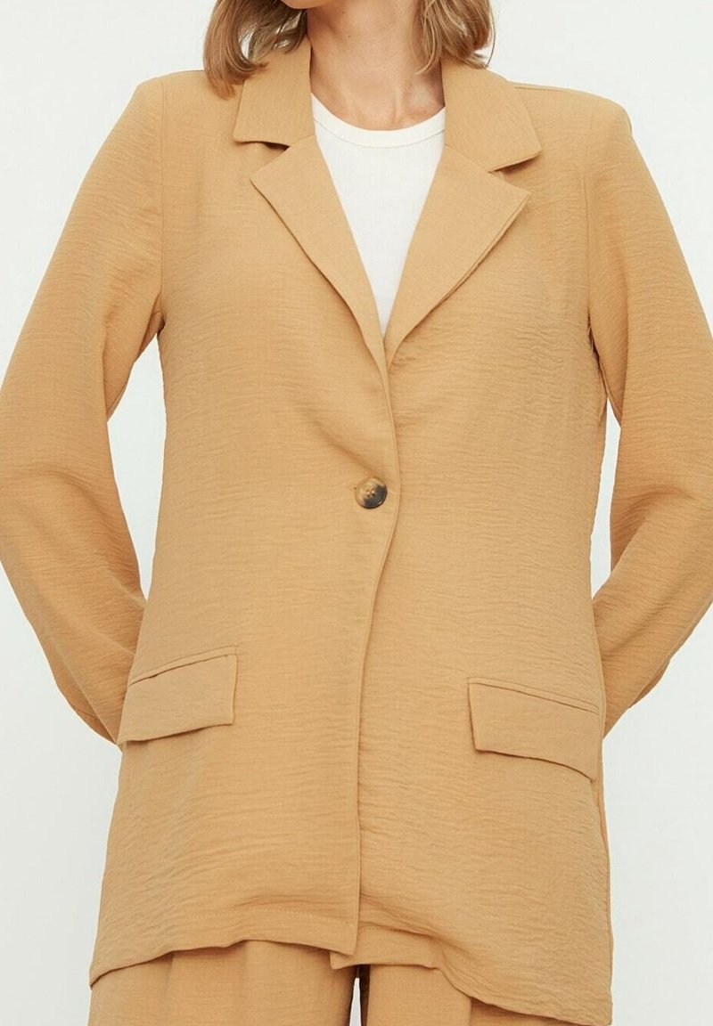 Blazer color marrón claro hecho de tejido texturizado, con solapa en pico, cierre de un solo botón y dos bolsillos frontales.
