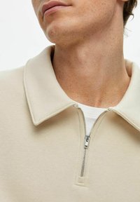 Närbild av en person som bär en beige zip-up skjorta med krage över en vit undertröja, vilket visar hals, haka och del av ansiktet.